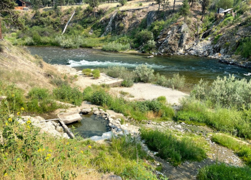 A serene river flows through a rocky landscape, surrounded by lush greenery and gentle hills under a clear sky.